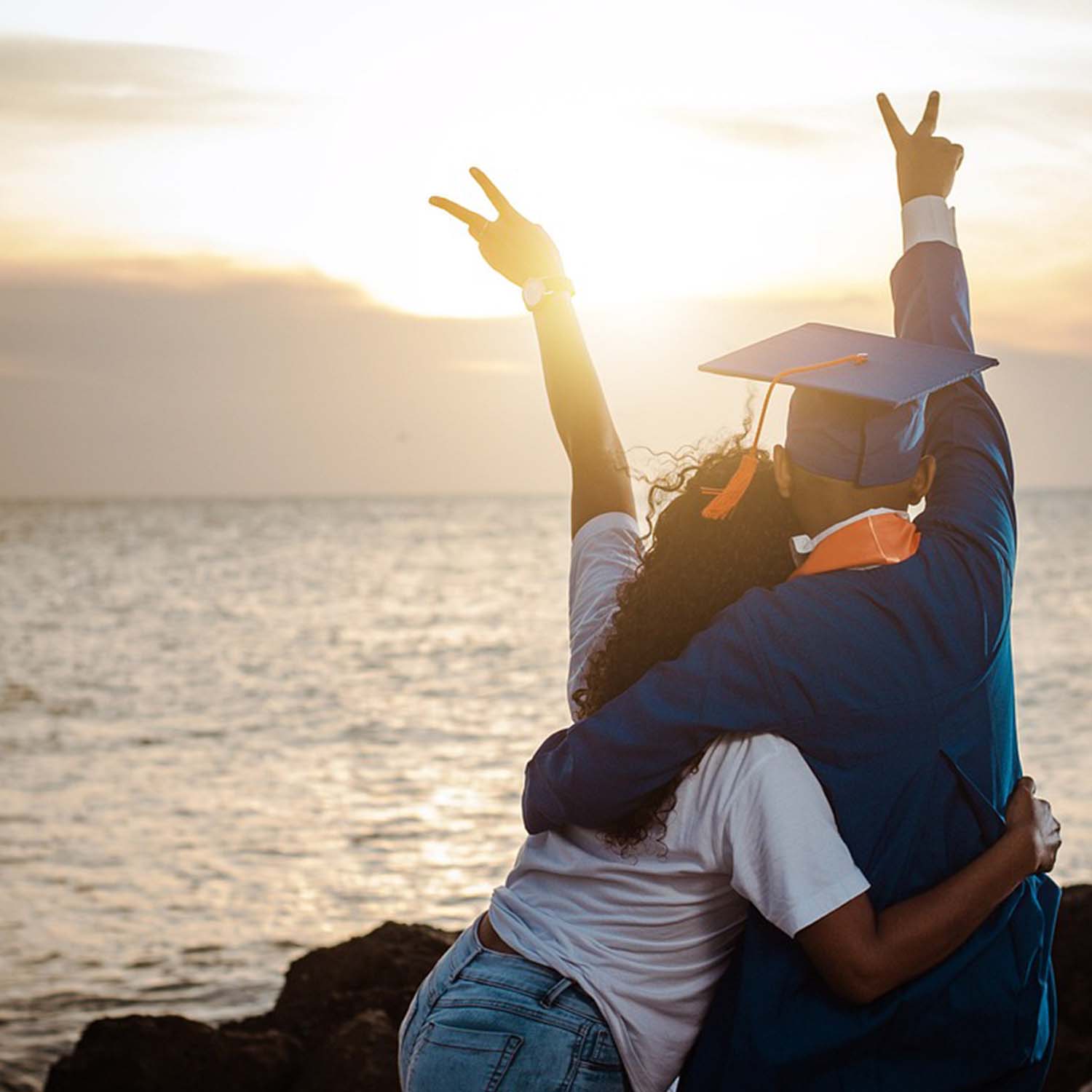 Photo of a person hugging someone in a graduation cap and gown while looking at the sunset.